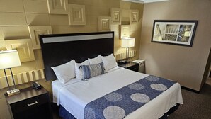 A hotel room with a large bed, a nightstand with a lamp, a dresser, and a carpeted floor. - The Landmark Hotel (Pikeville)
