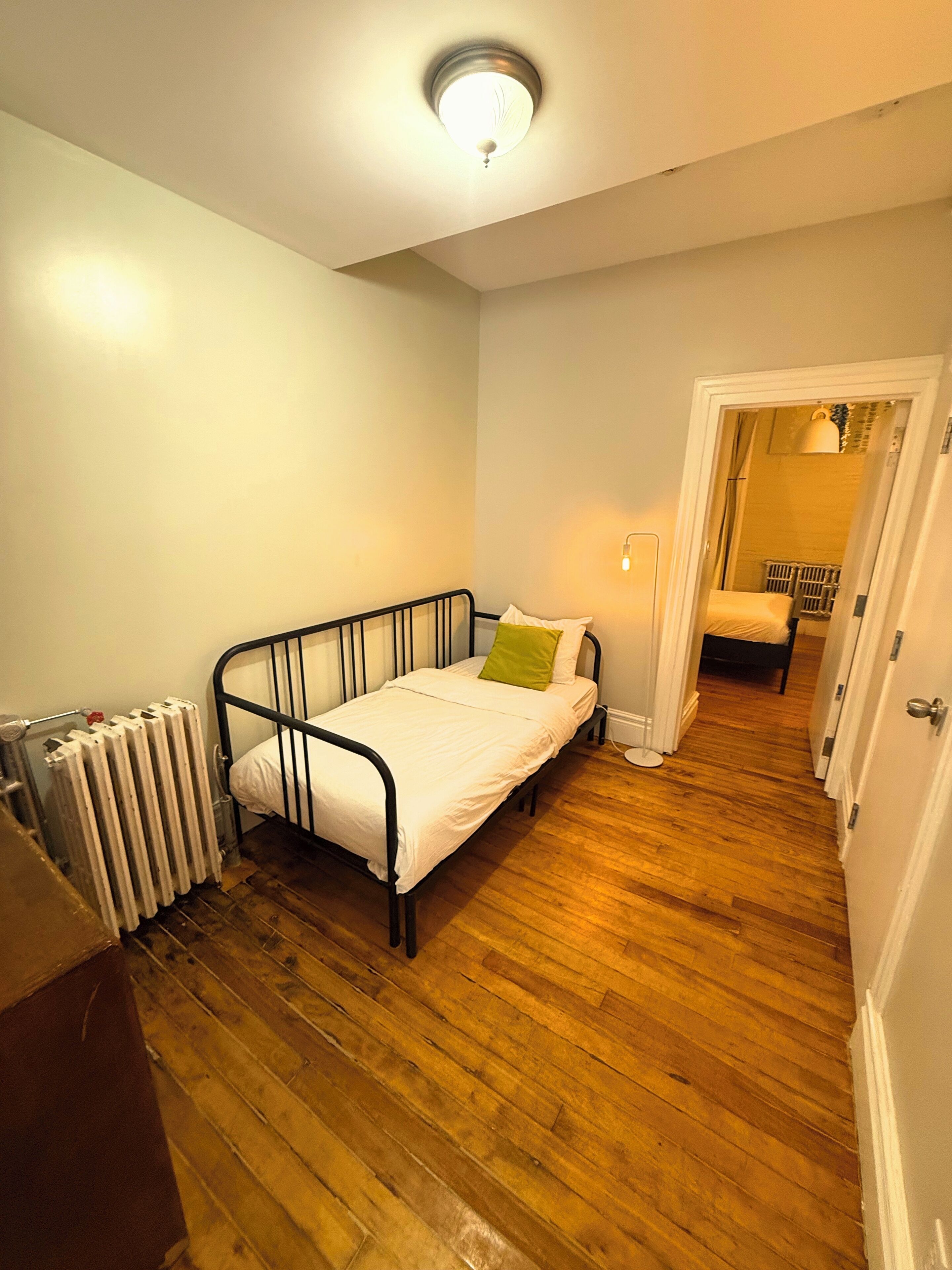 A narrow room with a single bed, a radiator, and a door leading to another room.