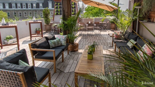 Un patio en la azotea con mobiliario de madera, plantas en macetas y vistas de edificios al fondo.