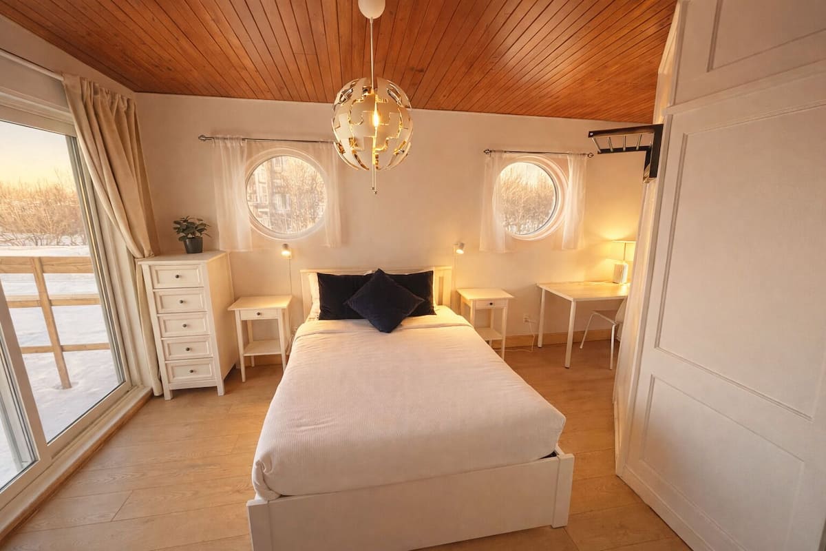 A bedroom with a bed, two bedside tables, a dresser, a window with curtains, and a wooden ceiling.