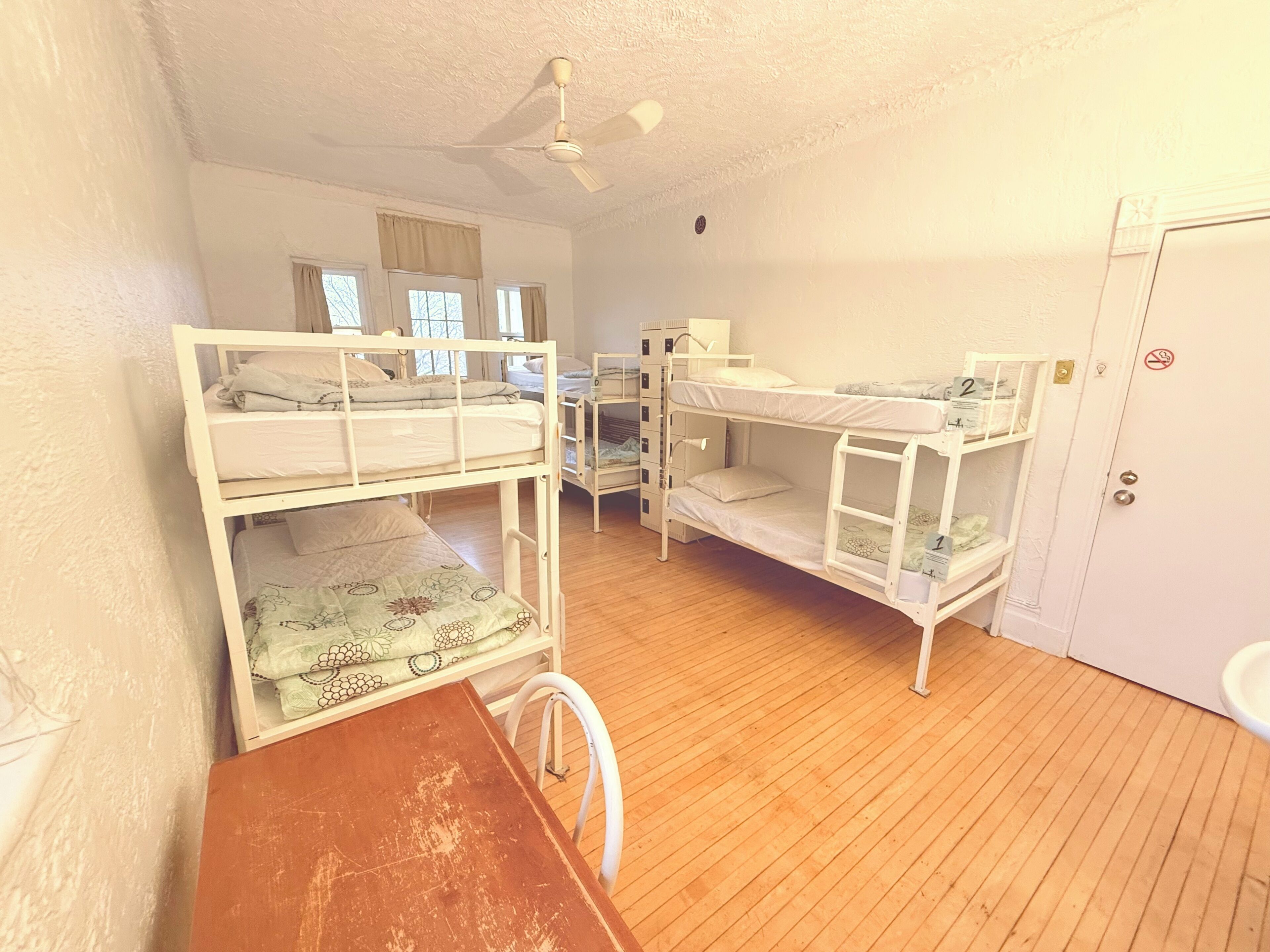 A room with bunk beds, a desk, a chair, a ceiling fan, and a door with a number.