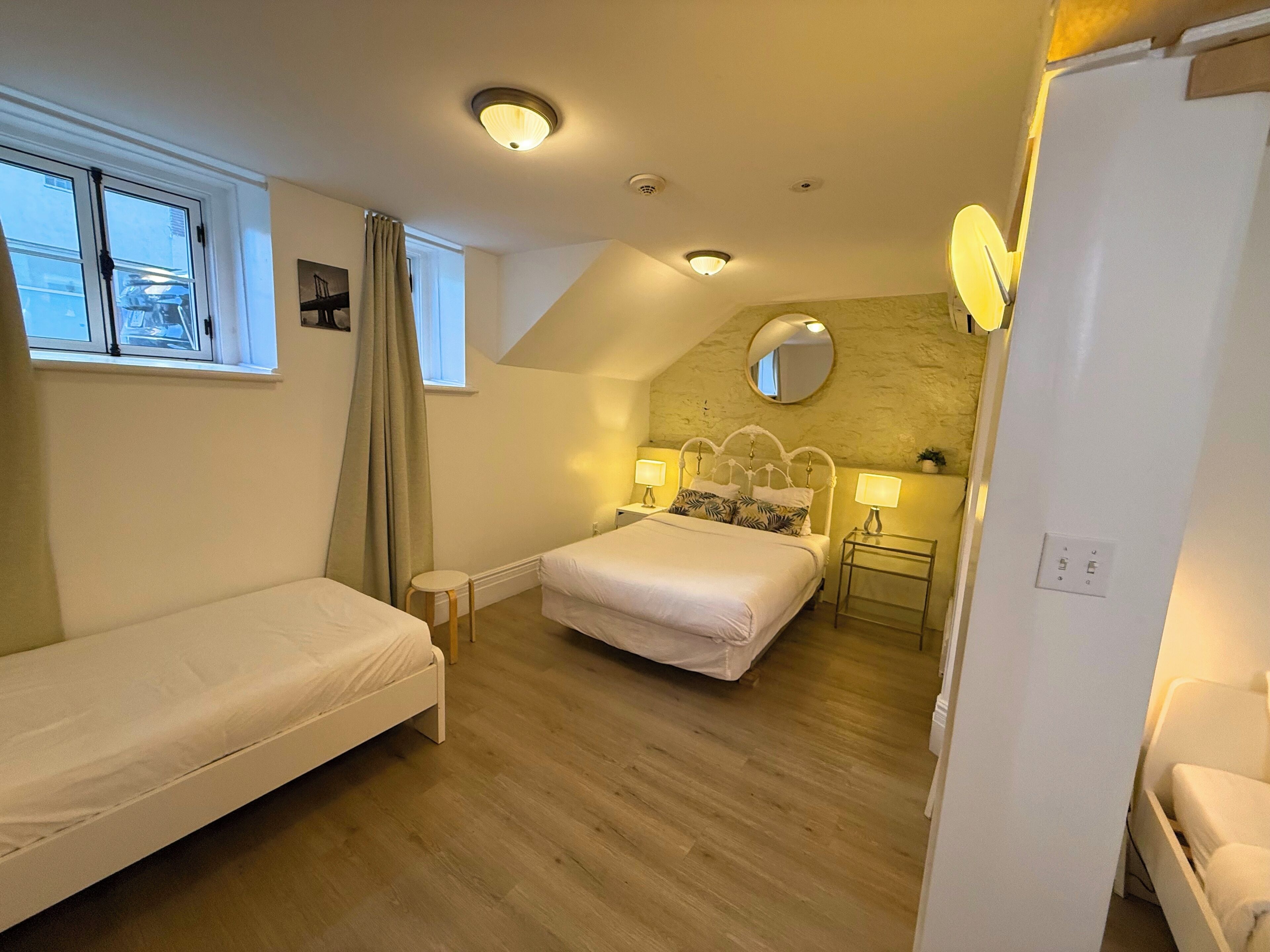 Un dormitorio con dos camas individuales, una mesita de noche pequeña, un velador, un espejo y una ventana con cortinas.