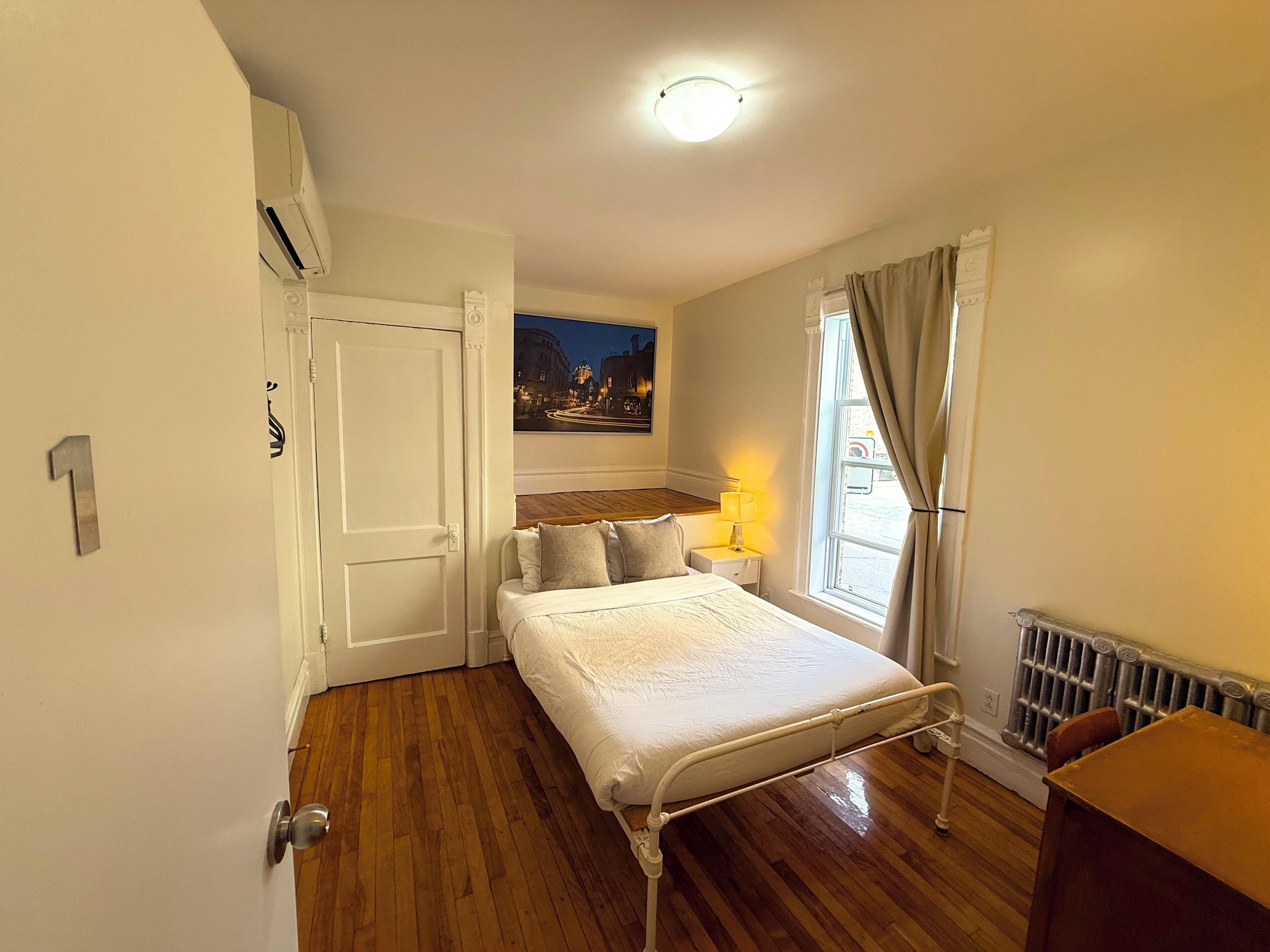 A bedroom with a bed, a nightstand, a window with curtains, a radiator, and a door.