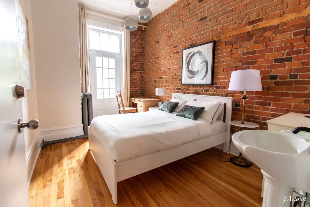 A modern hotel room with a brick wall, a large bed, a bedside table, a lamp, and a sink.