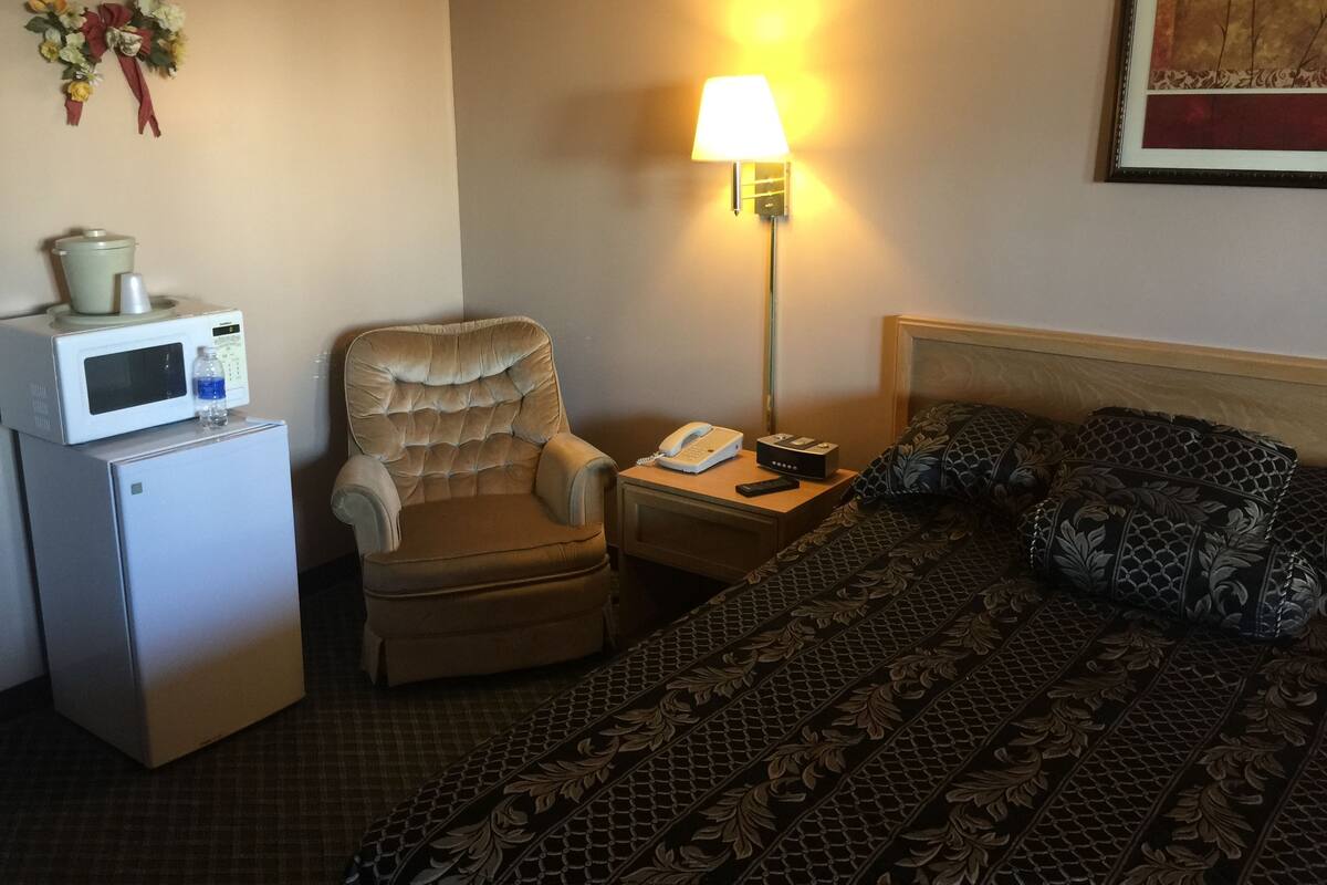 A hotel room with a bed, a chair, a nightstand with a lamp, a microwave, and a refrigerator.