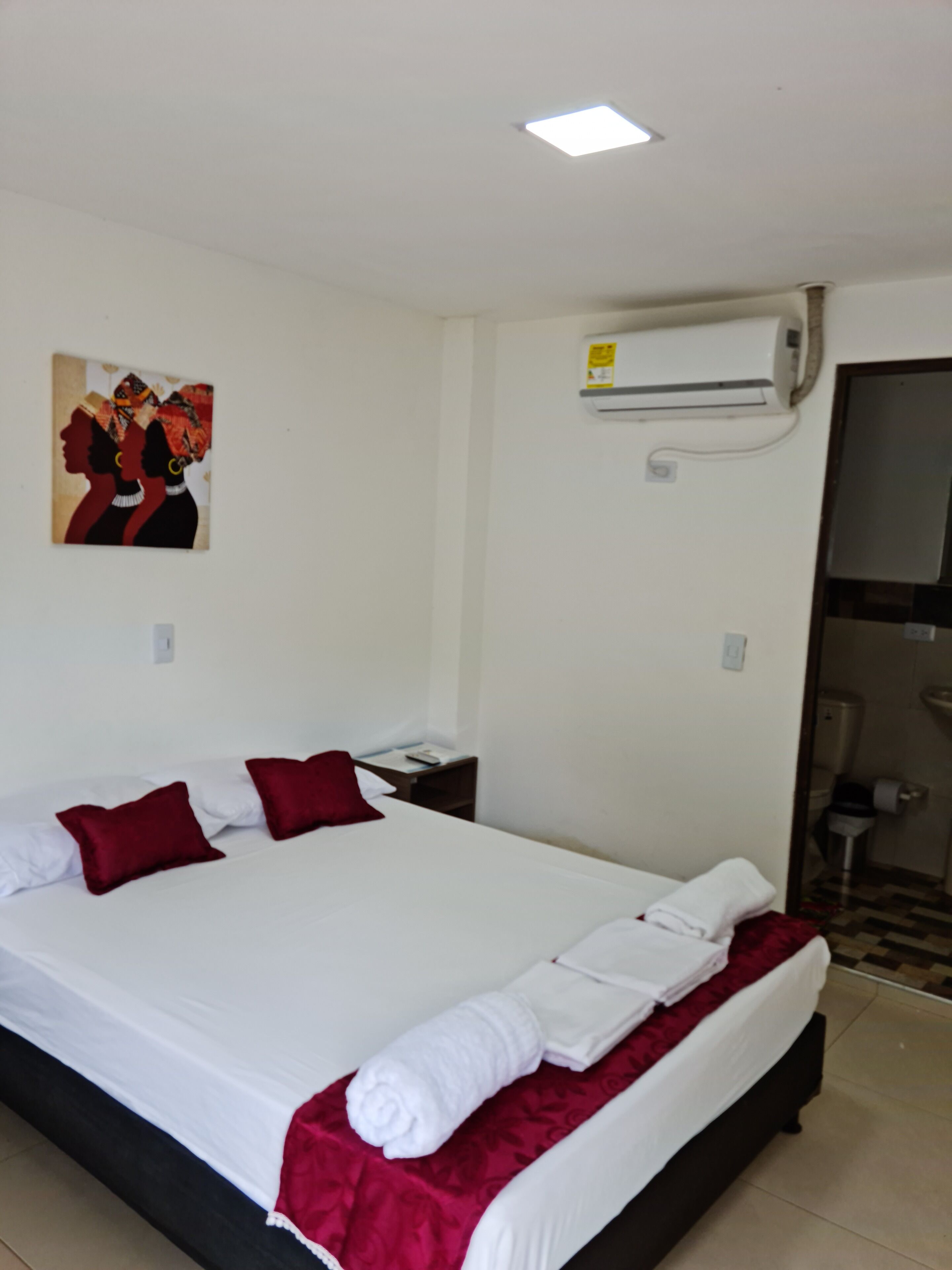 A hotel room with a large bed, red and white pillows, a white blanket, and a red throw. There is a painting on the wall, an air conditioning unit, and a small table with a remote control.