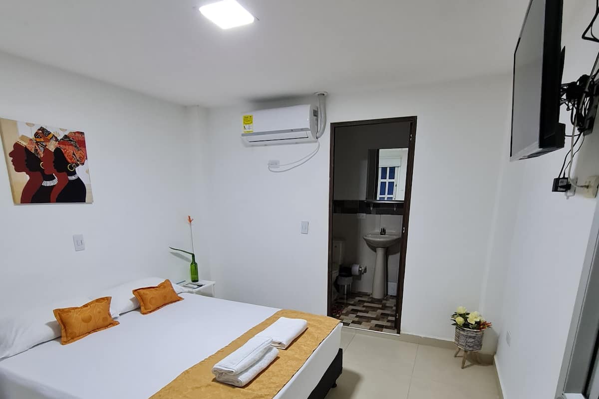 Ein Hotelzimmer mit Bett, Nachttisch, Wandklimaanlage, einem durchgegangenen Tür sichtbaren Badezimmer und einem an der Wand befestigten Fernseher.