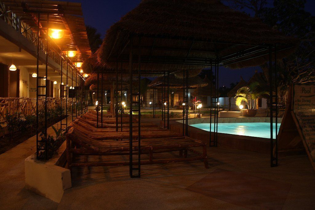 Un espace piscine agrémenté de bancs en bois et d’une structure à toit de chaume, entouré d’un bâtiment doté de nombreux balcons et éclairages.