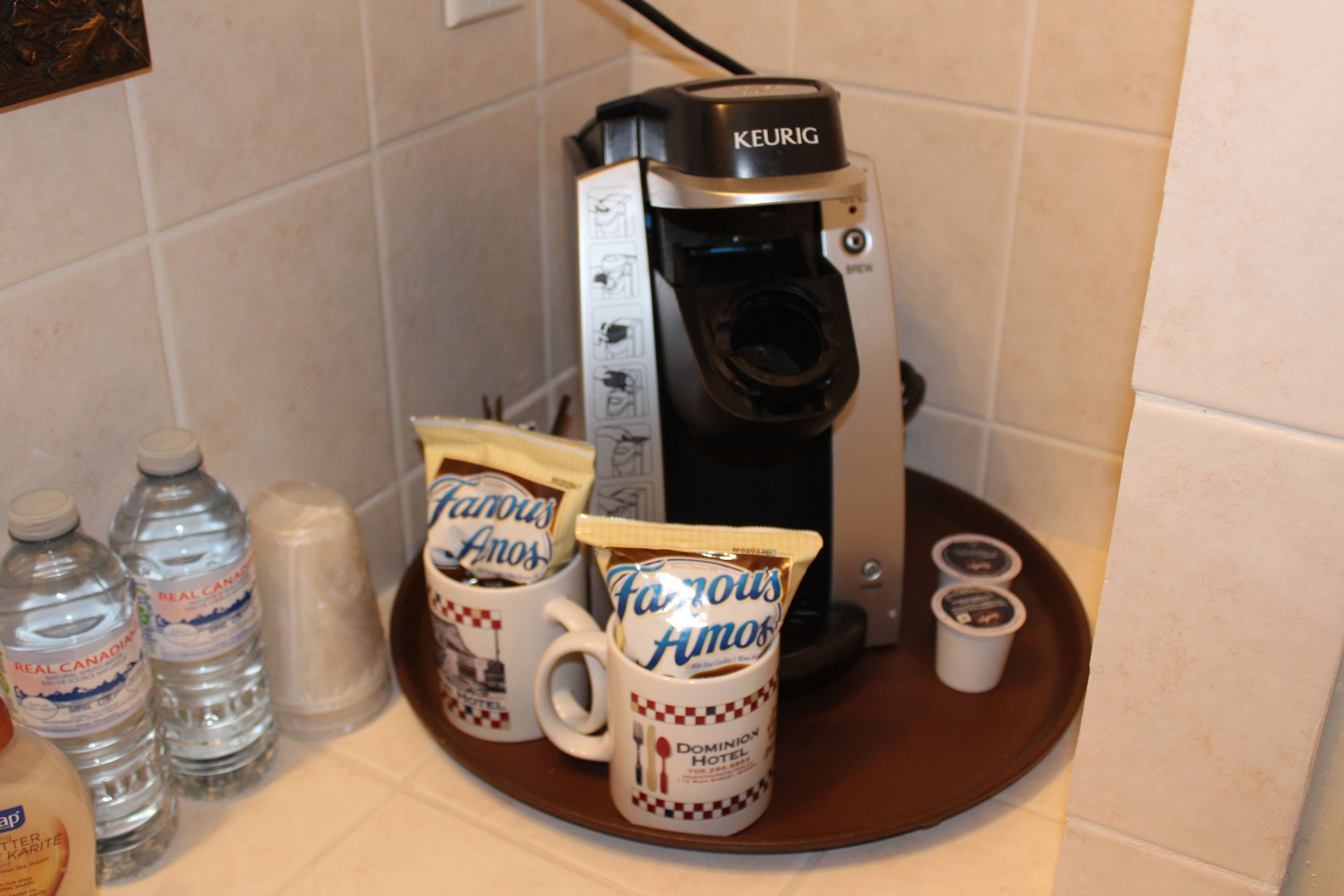 A Keurig coffee maker on a tray with two mugs labeled 'Famous Anos' and a bottle of water.