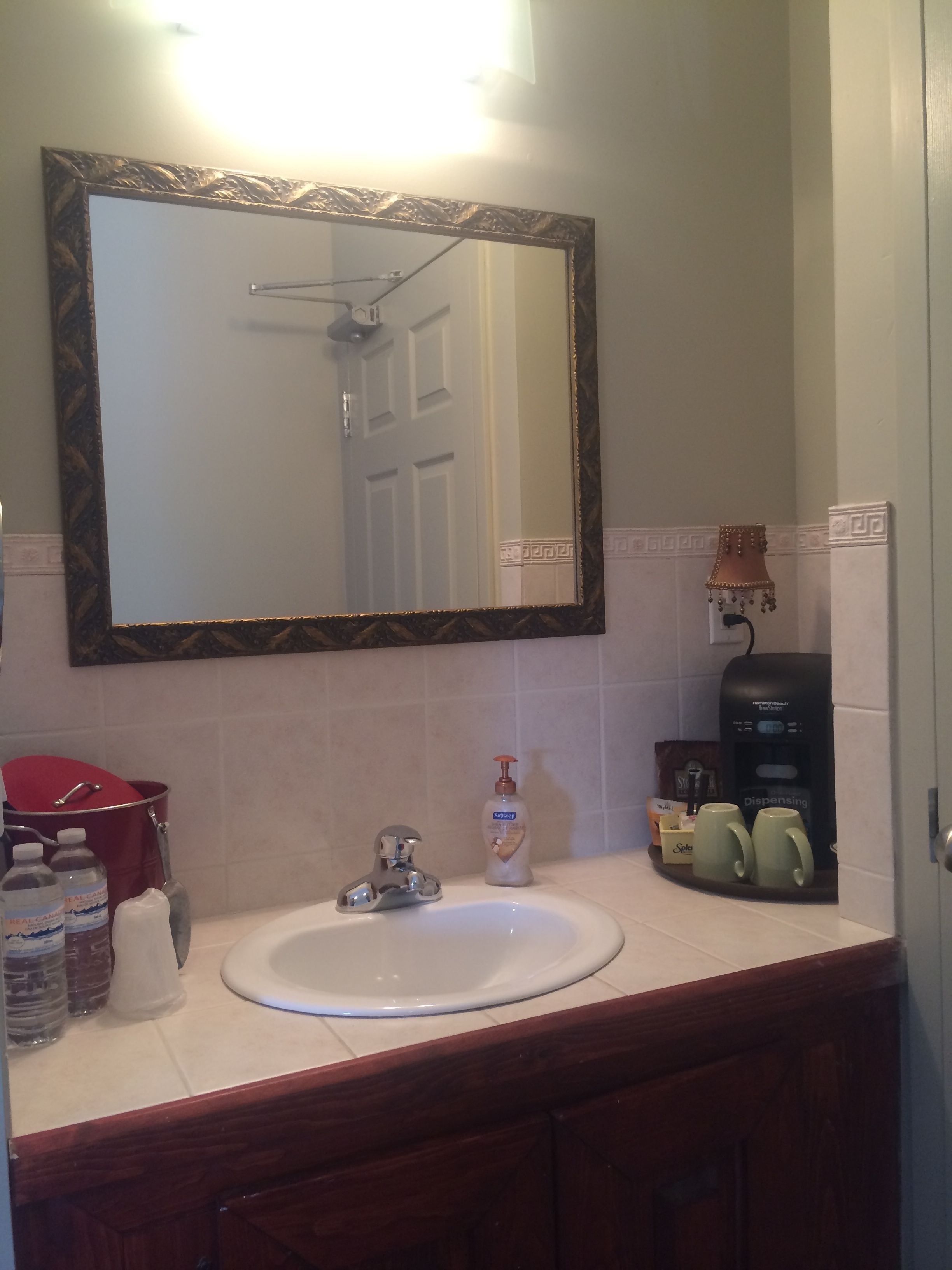 A bathroom with a white sink, a mirror with a decorative frame, a red bucket, a soap dispenser, and a coffee machine.