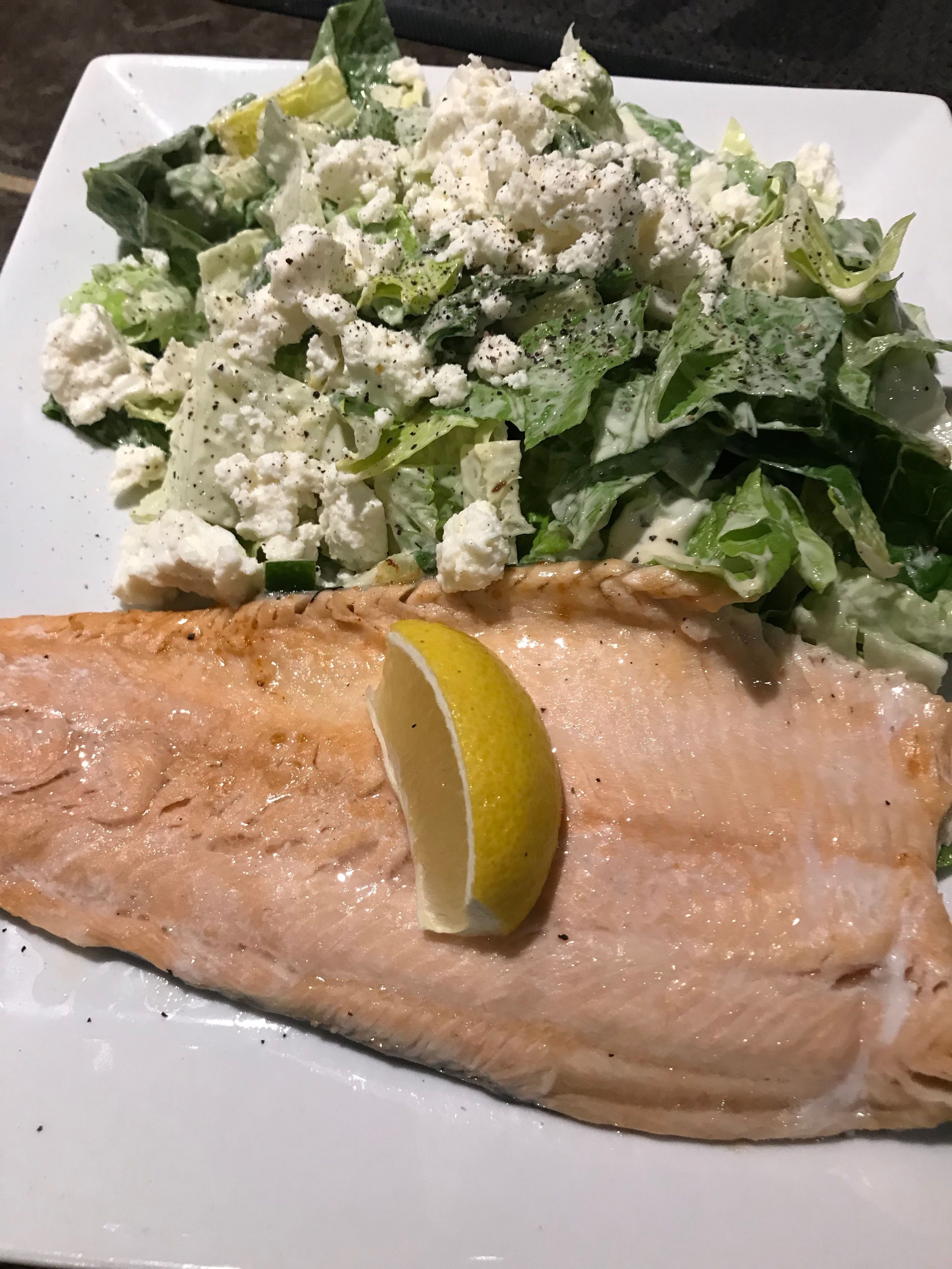 A plate with a piece of salmon, a lemon wedge, and a salad with feta cheese.