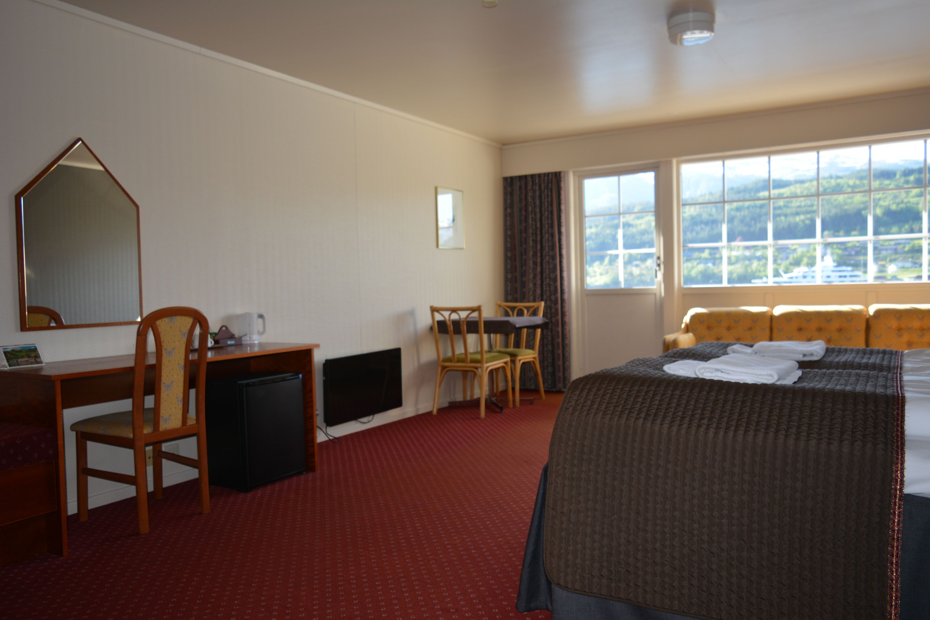 A hotel room with a bed, a desk, a chair, a small refrigerator, and a window with a view of the outdoors.