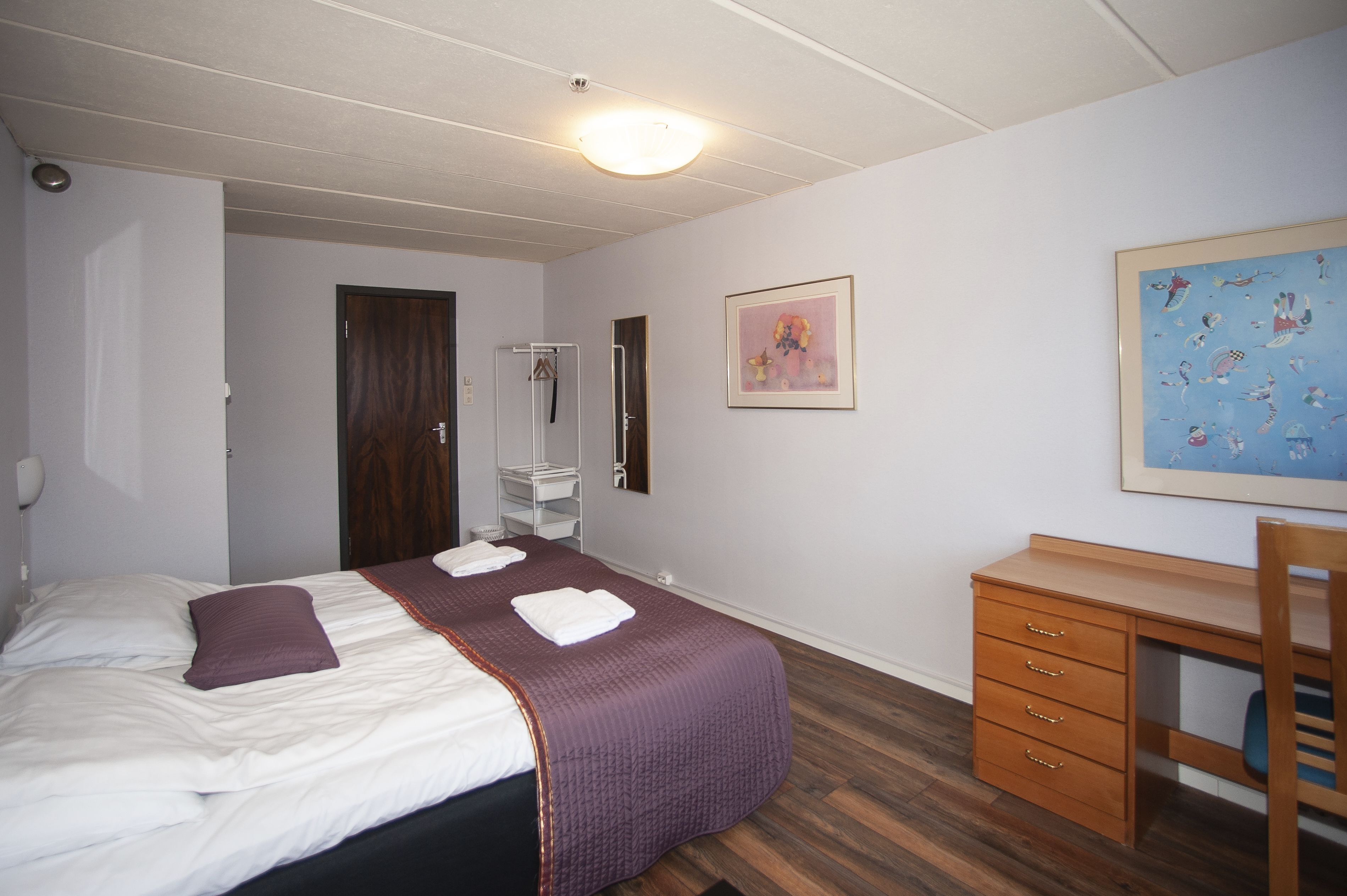 A bedroom with a bed, a desk, a chair, a wardrobe, and a painting on the wall.