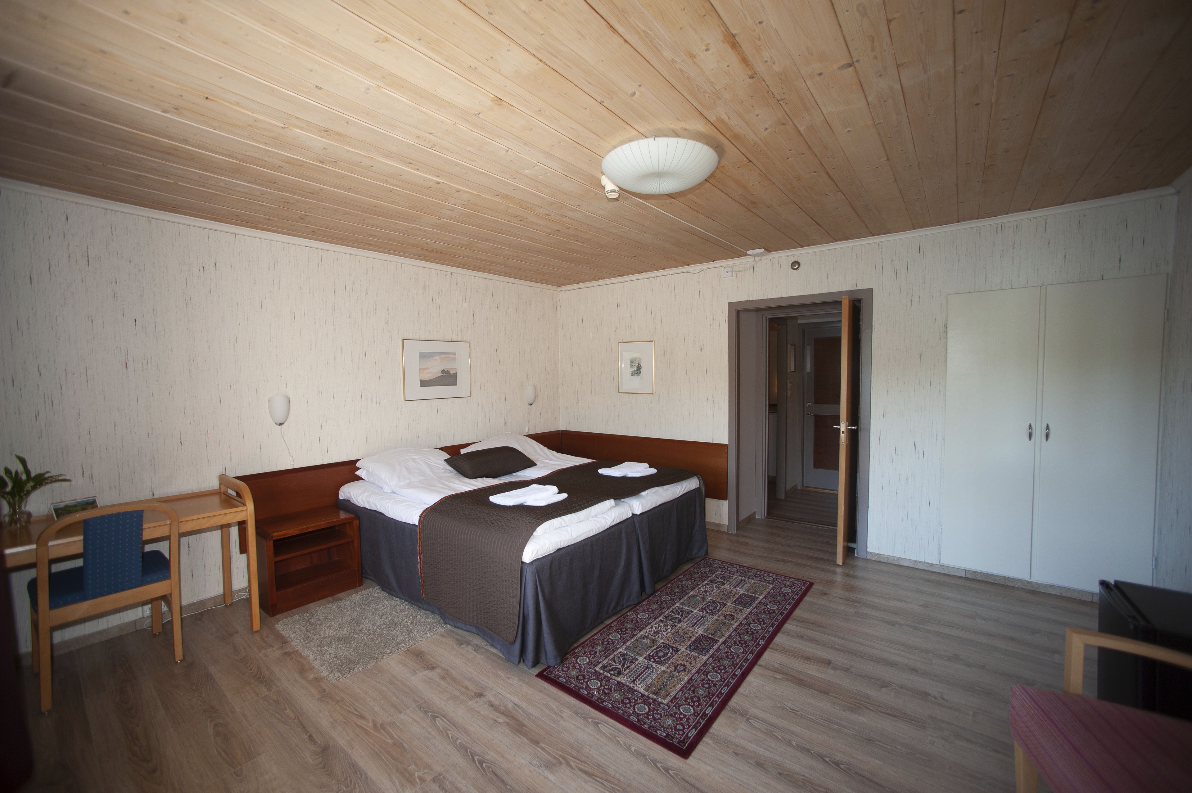 A bedroom with a wooden ceiling, a bed with a grey and brown bedspread, a wooden desk with a chair, and a wardrobe.