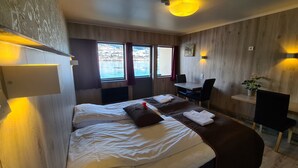A hotel room with a bed, a desk, and a chair. There are windows with curtains, a plant, and a wall-mounted light fixture. - Strand Fjordhotel (Ulvik)
