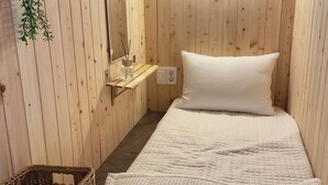 Una habitación pequeña y acogedora con paredes de madera, una cama individual con ropa blanca, un estante con un espejo y un candelabro, y una cesta de mimbre.