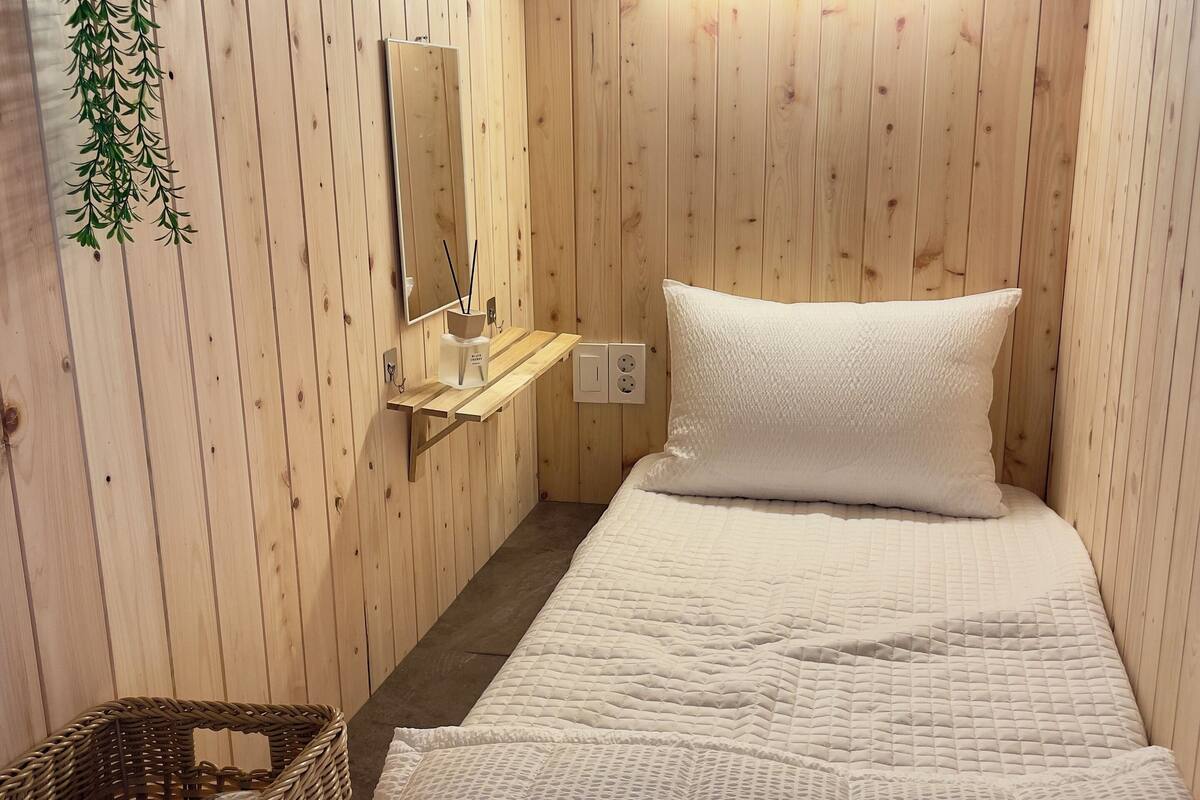 A small, cozy room with wooden walls, a single bed with white bedding, a small shelf with a mirror and a candle holder, and a wicker basket.