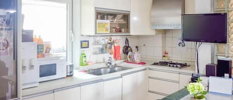 Una cocina moderna con armarios blancos, un microondas, un fregadero y una estufa. Hay un televisor montado en la pared y una pequeña mesa con un florero y una taza.