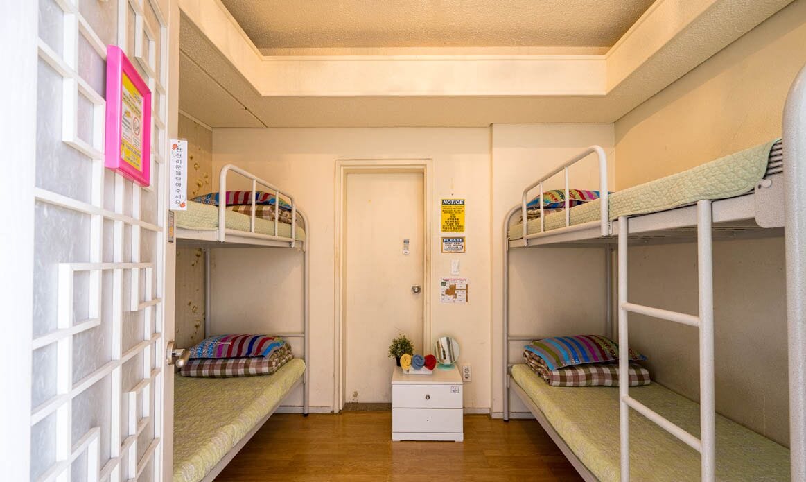 Une chambre de dortoir avec des lits superposés, une petite commode blanche et une porte sur laquelle est affichée une annonce.