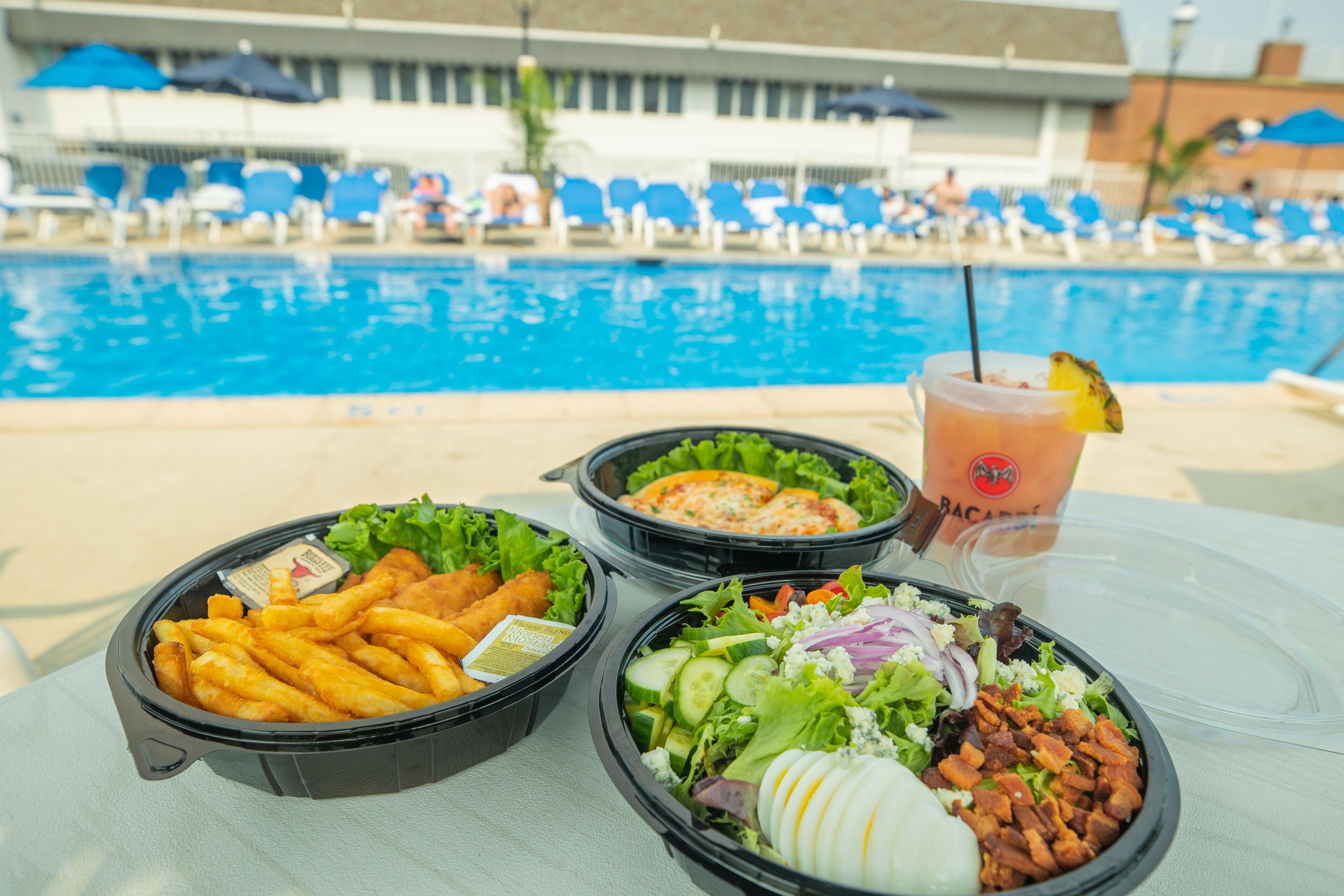 Una mesa con recipientes de comida y una bebida cerca de una piscina, con sillas de descanso y sombrillas.