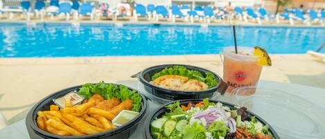Una mesa con recipientes de comida y una bebida cerca de una piscina, con sillas de descanso y sombrillas.