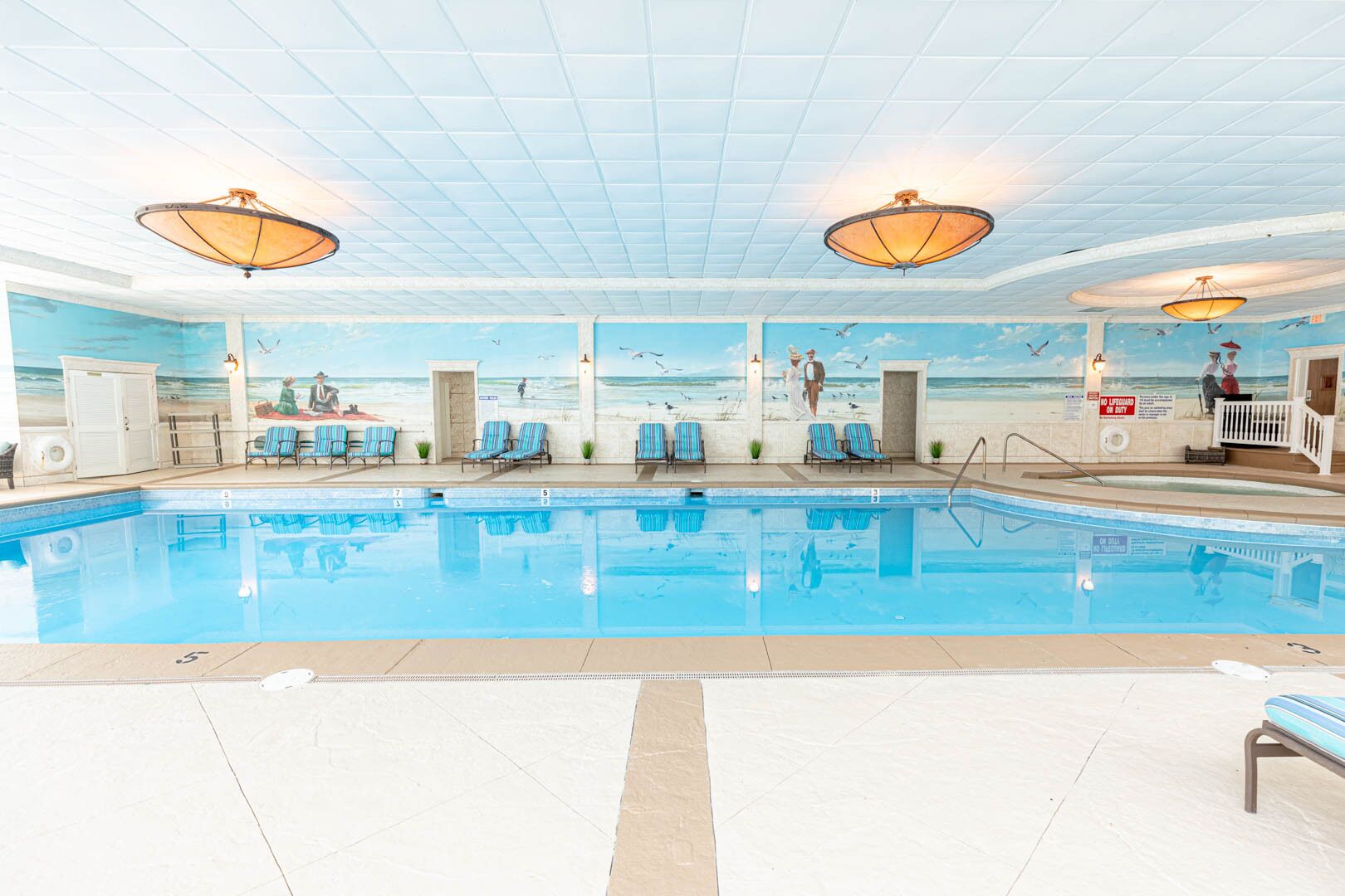 An indoor swimming pool with lounge chairs, a lifeguard station, and murals of beach scenes.
