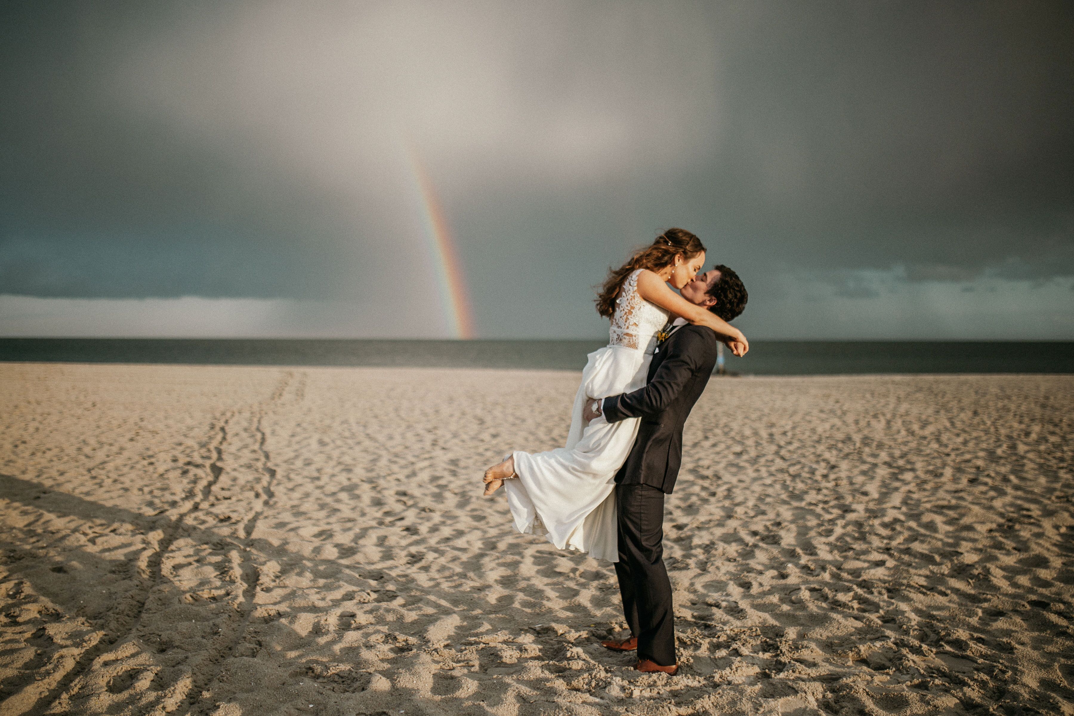 Een stel omarmt elkaar op het strand, met een regenboog op de achtergrond.