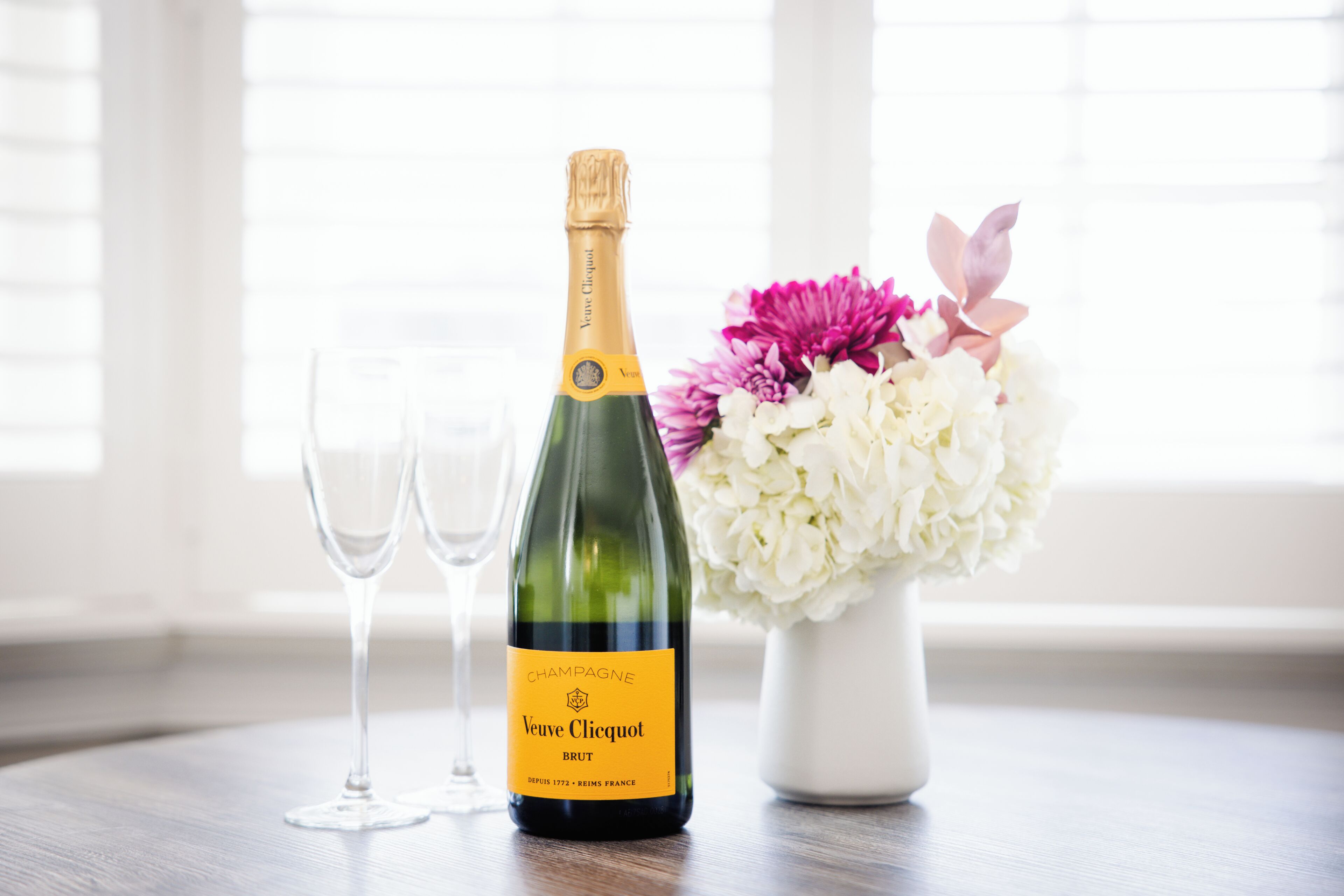 A bottle of Veuve Clicquot Brut champagne, two empty glasses, and a vase with flowers on a table.