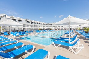 A poolside area with blue lounge chairs and white umbrellas, a clear blue sky, and a building with balconies in the background.