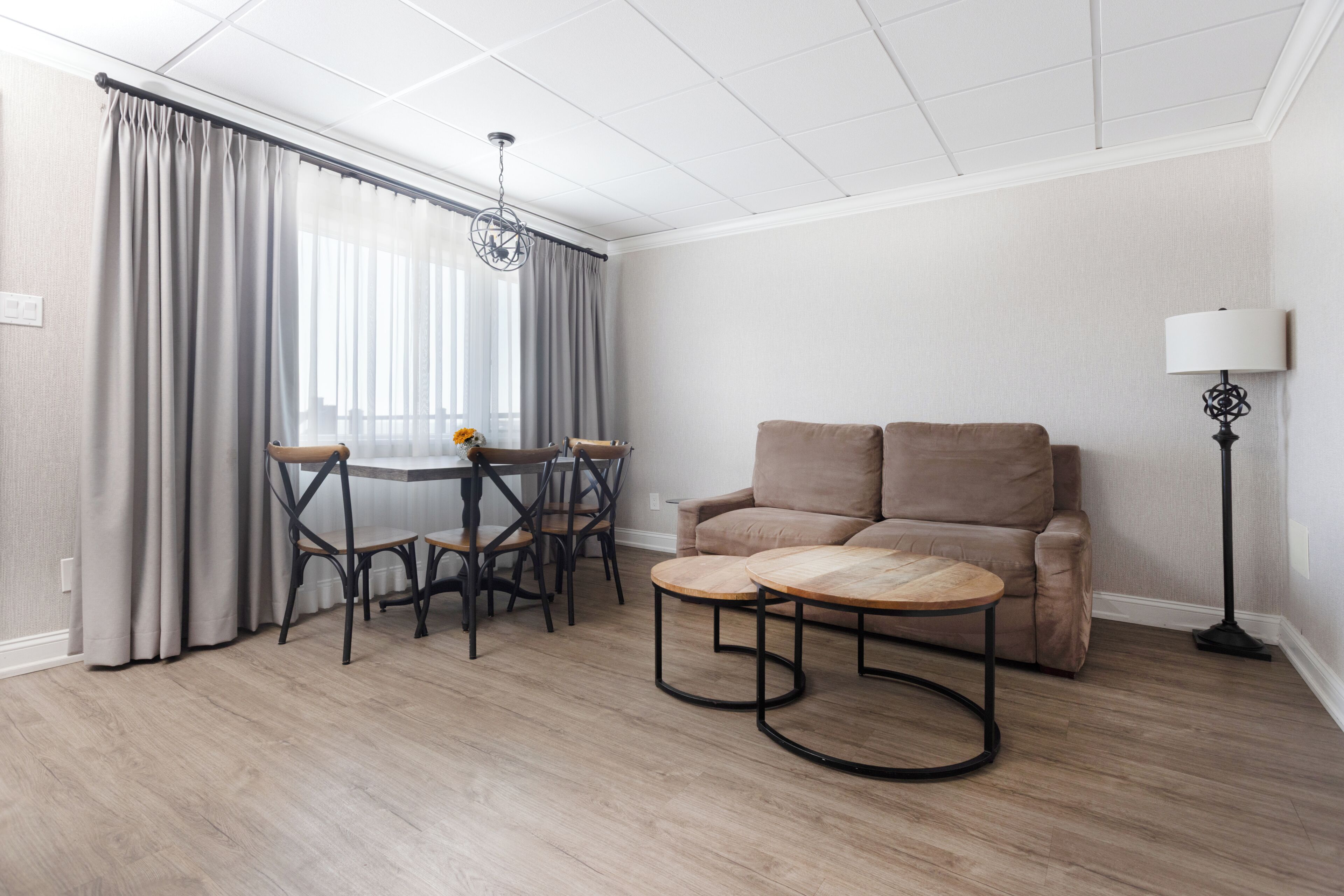 A room with a dining table, chairs, a sofa, a lamp, and a window with curtains.
