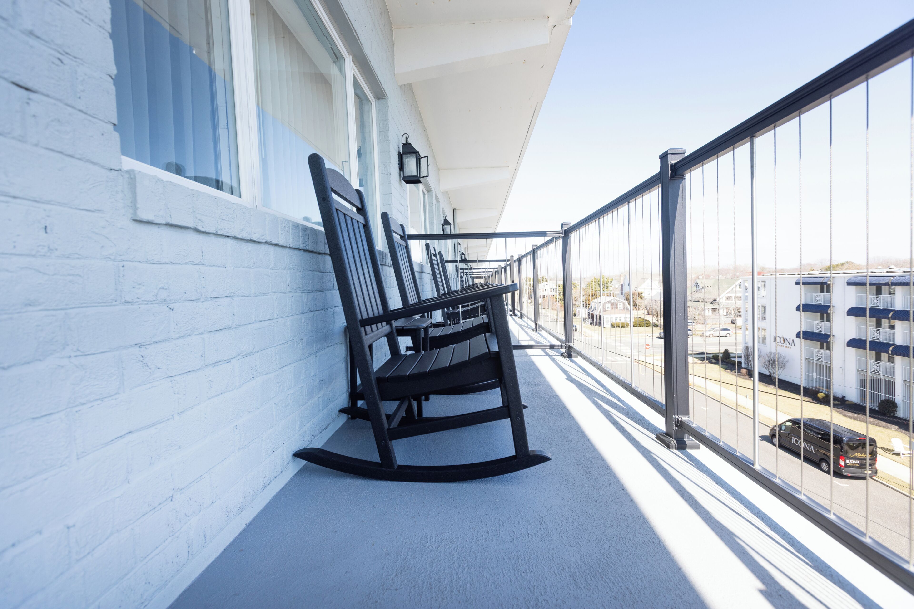 A balcony with a railing, a row of black rocking chairs, and a view of a street with parked cars and buildings.