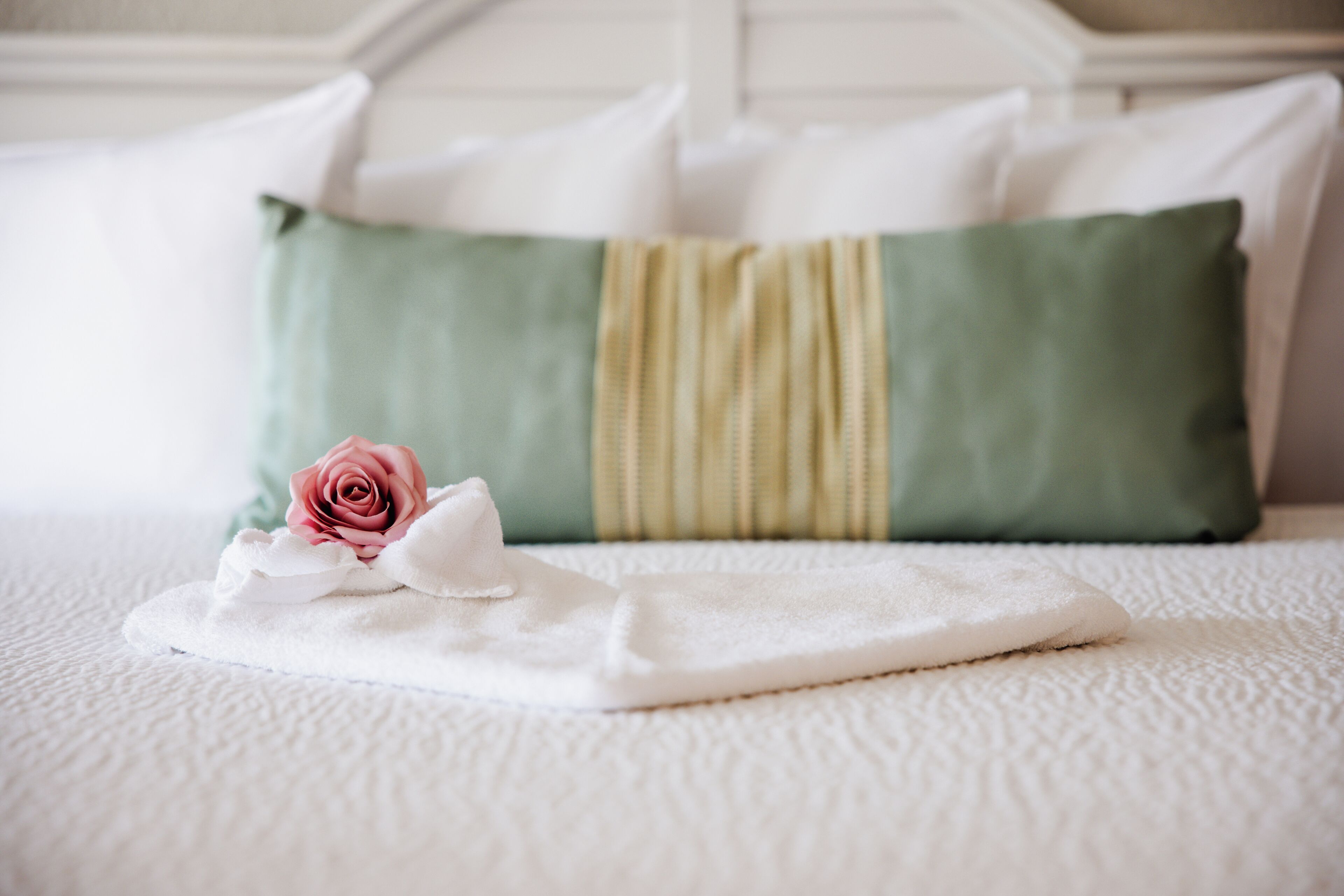Una cama bien hecha con una almohada verde y blanca, una toalla blanca y una rosa rosa.