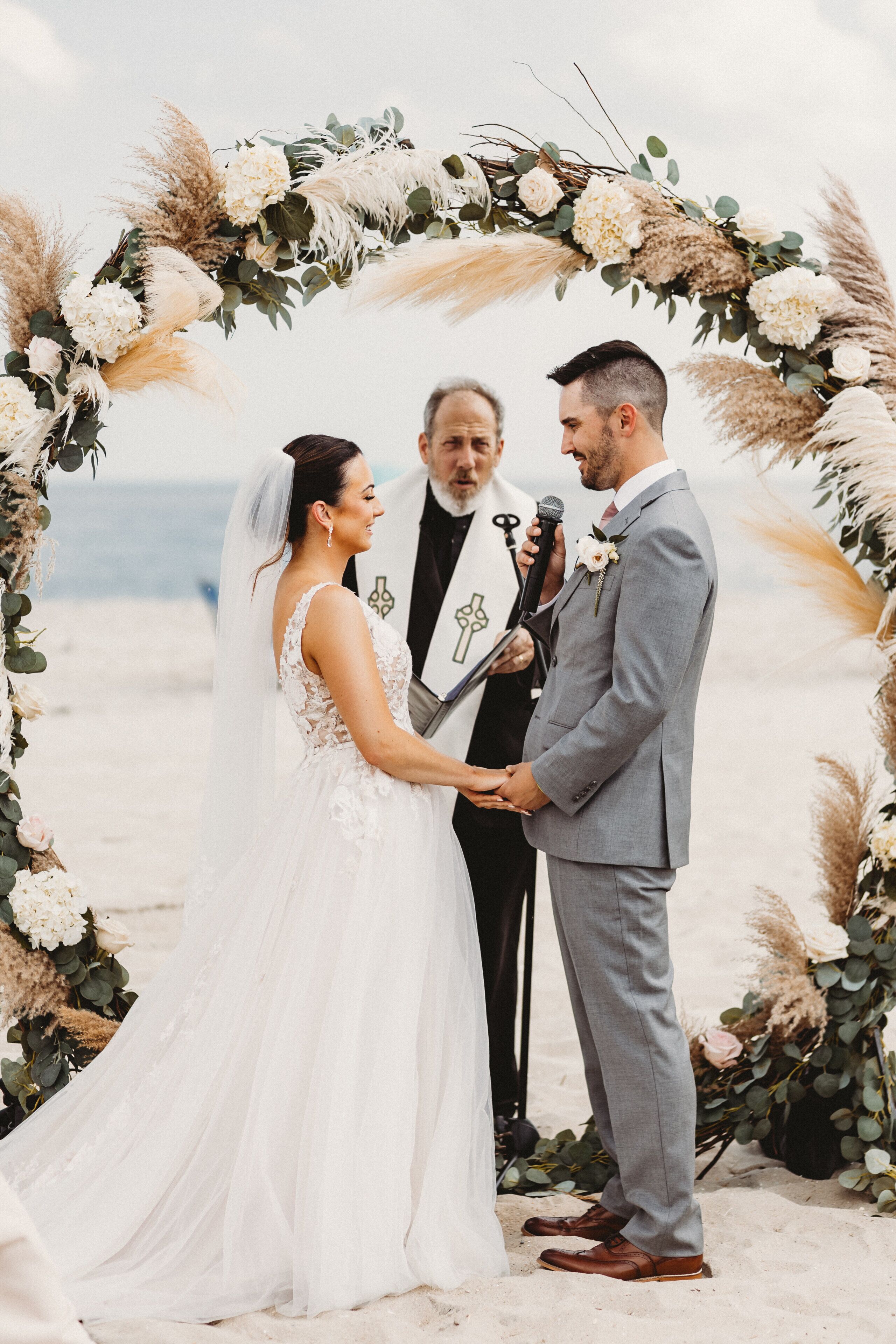 A wedding ceremony on a beach with a floral arch, officiant, and bride and groom.
