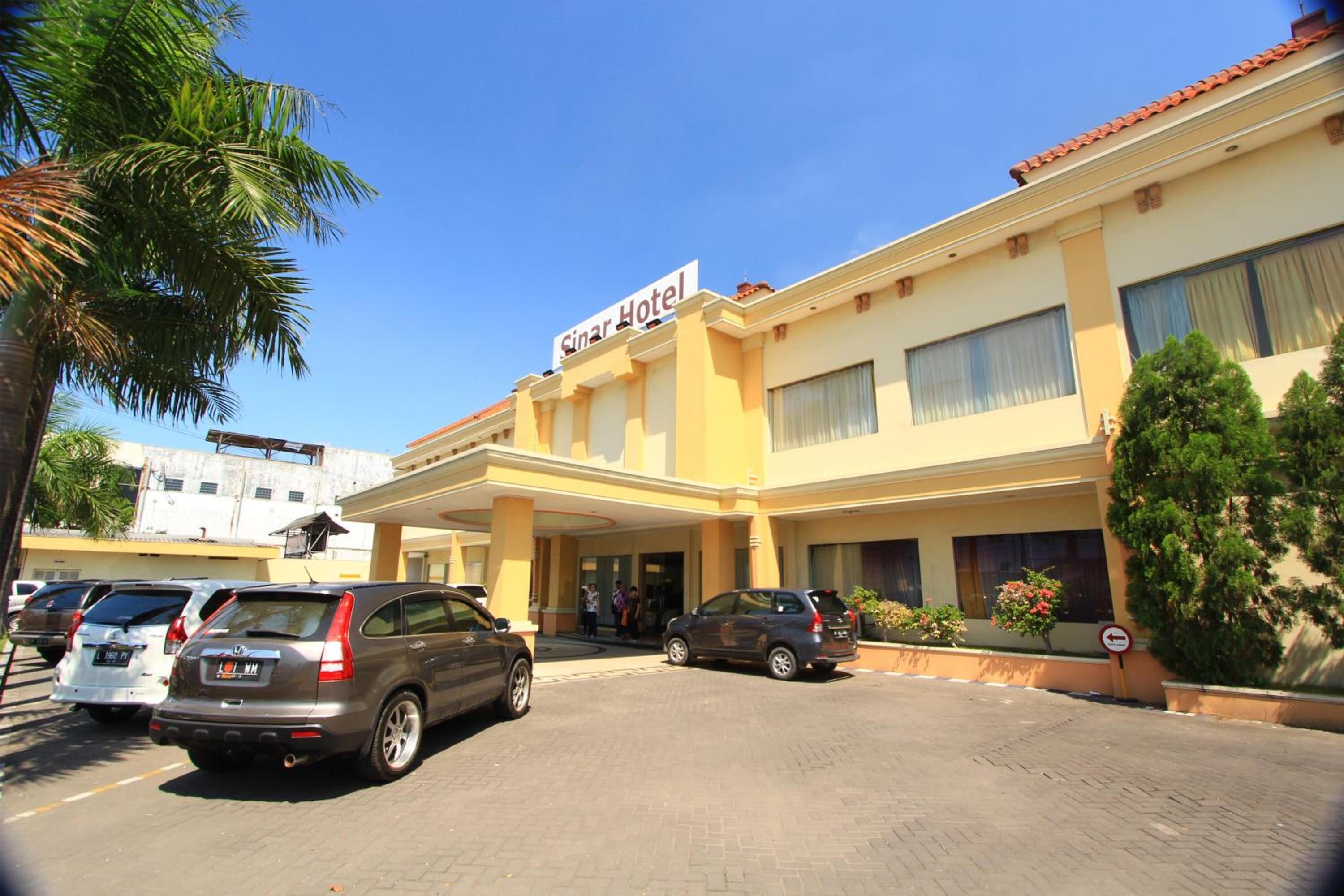 A hotel with a sign reading 'Sinar Hotel,' featuring a modern facade, a covered entrance, and a parking area with several cars.
