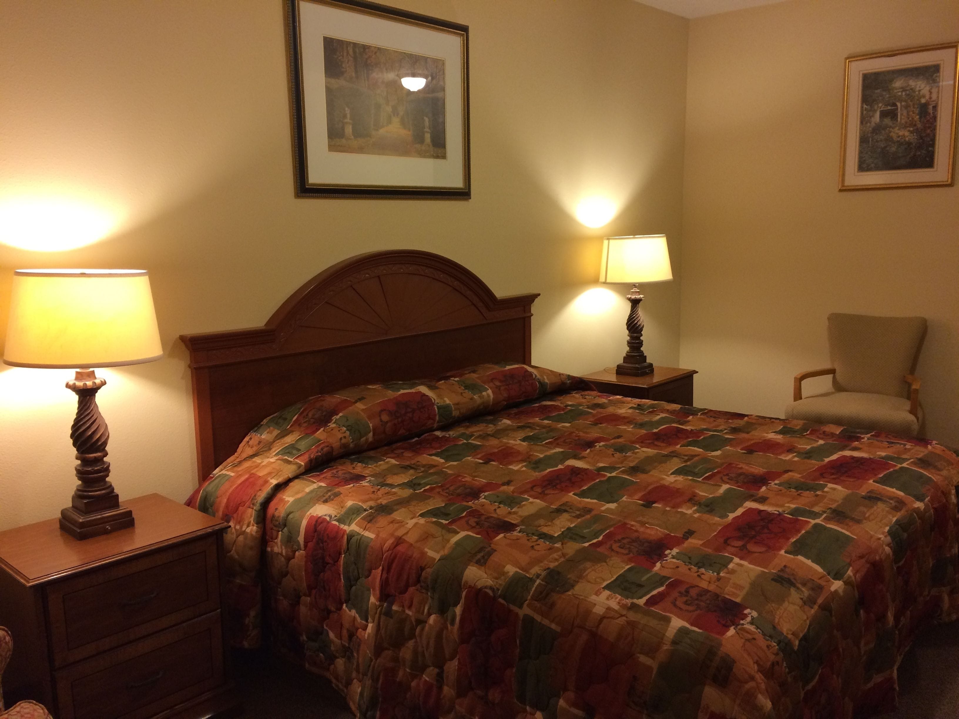 A neatly made bed with a quilt, two bedside tables with lamps, a chair, and two framed pictures on the walls.