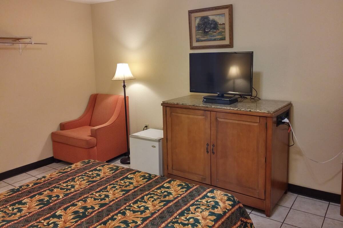 Hotellihuone, jossa on sänky, puinen kaappi, televisio, lamppu ja taulu seinällä.