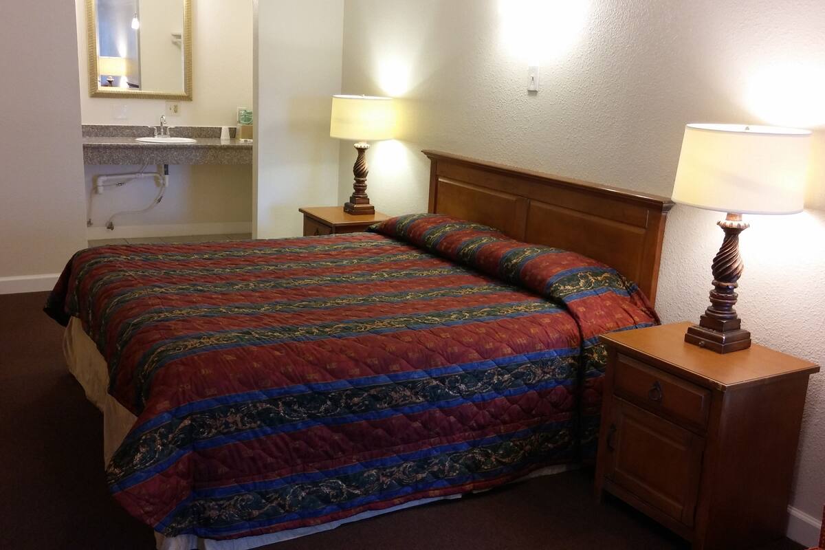 Hotellihuone, jossa on suuri sänky, kaksi yöpöytää valaisimineen, puinen sängynpääty, pieni yöpöytä valaisimella ja seinälle kiinnitetty peili.