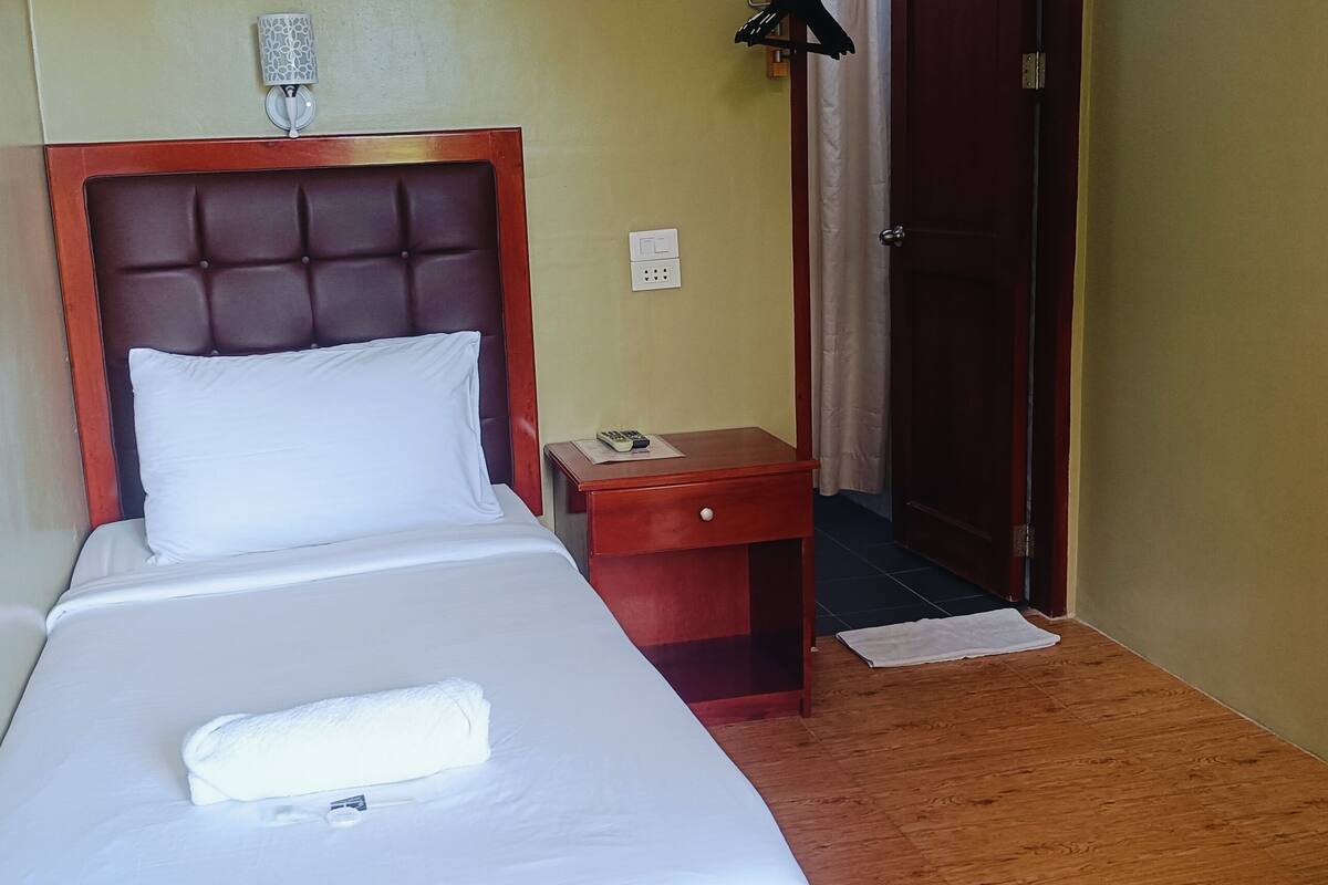 A neatly made bed with a white pillow and a folded towel, a wooden nightstand with a lamp, and a door leading to another room.