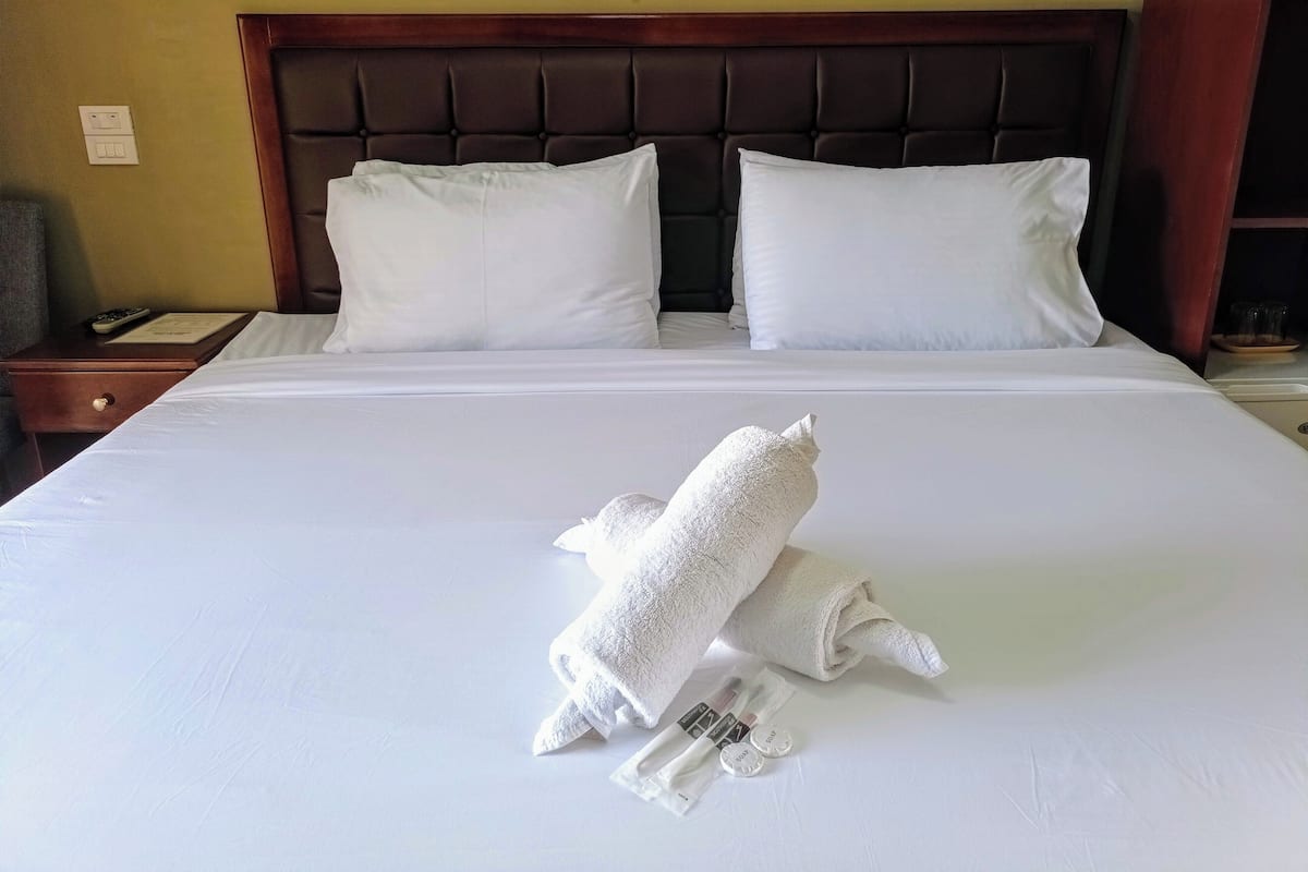 A neatly made bed with white linens, a folded towel, and toiletries on the bed.