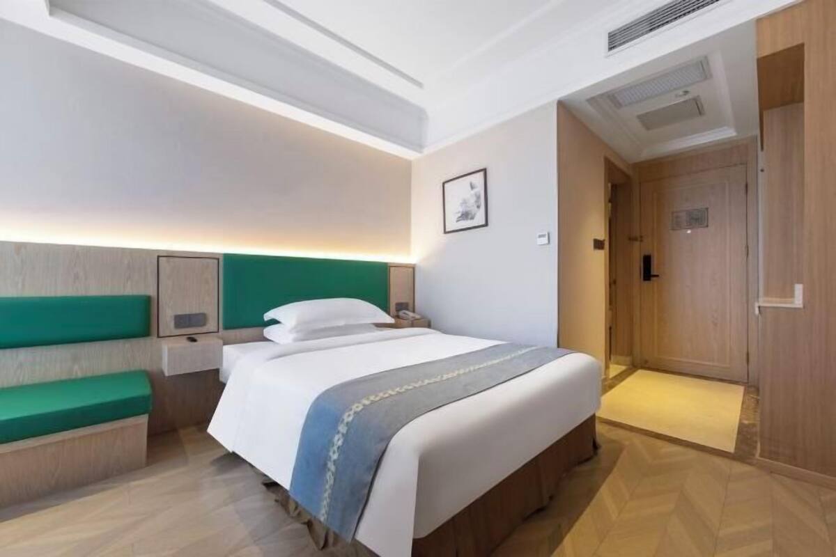 A hotel room with a bed, a bedside table, a green bench, a wall-mounted headboard, and a door leading to a bathroom.