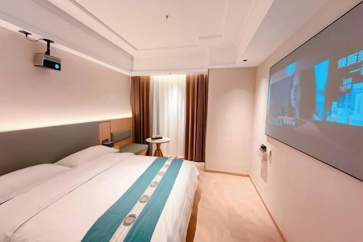 A hotel room with a bed, a projector screen, a small table, and a window with curtains.