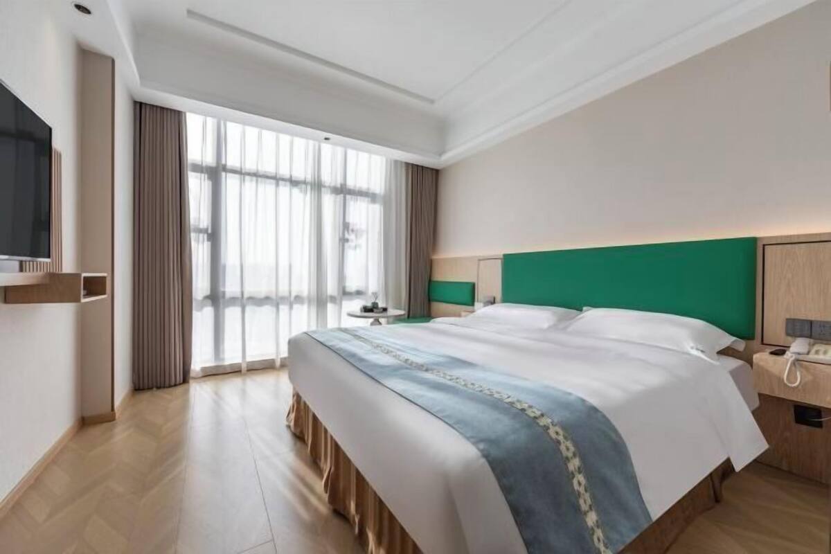 A hotel room with a large bed, a bedside table with a lamp, a TV mounted on the wall, and a large window with curtains.