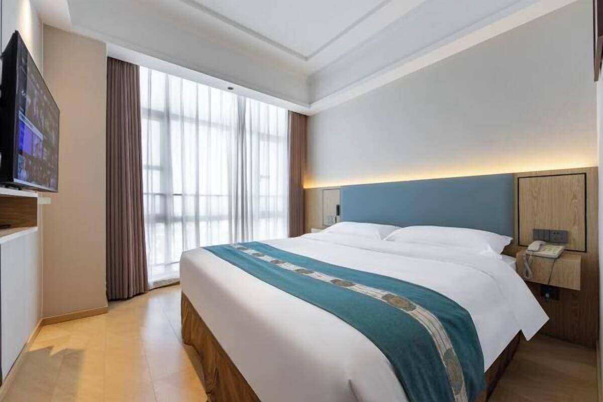 A hotel room with a large bed, a flat-screen TV, a bedside table with a lamp, and a large window with curtains.