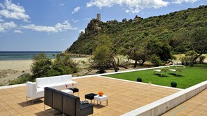 A terrace with outdoor seating, a view of the beach, and a hill with ruins.