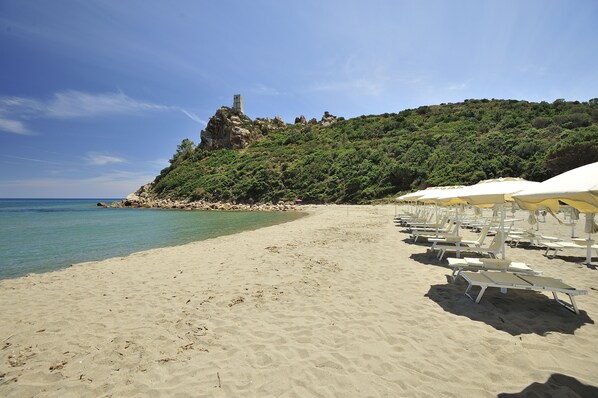 A beach with white lounge chairs and umbrellas, a rocky cliff with a tower, and clear blue water.