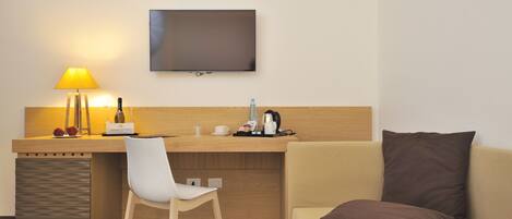 A hotel room with a wooden desk, a white chair, a brown sofa, a television, a lamp, and a coffee maker.