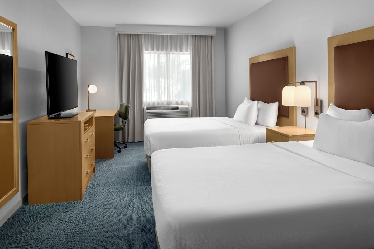 Hotellihuone, jossa on kaksi sänkyä, työpöytä, jossa on televisio, tuoli, lamppu ja ikkuna, jossa on verhot.