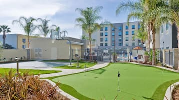 Campo de golf con putting green, rodeado de palmeras y un edificio con múltiples ventanales.