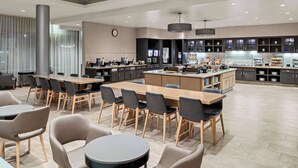 Un moderno vestíbulo de hotel con zona de comedor central, mostrador de recepción y un gran reloj en la pared.