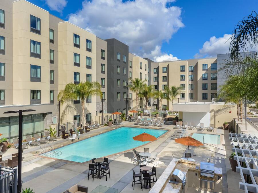 A hotel pool area with multiple lounge chairs and umbrellas, surrounded by a multi-story building with balconies.
